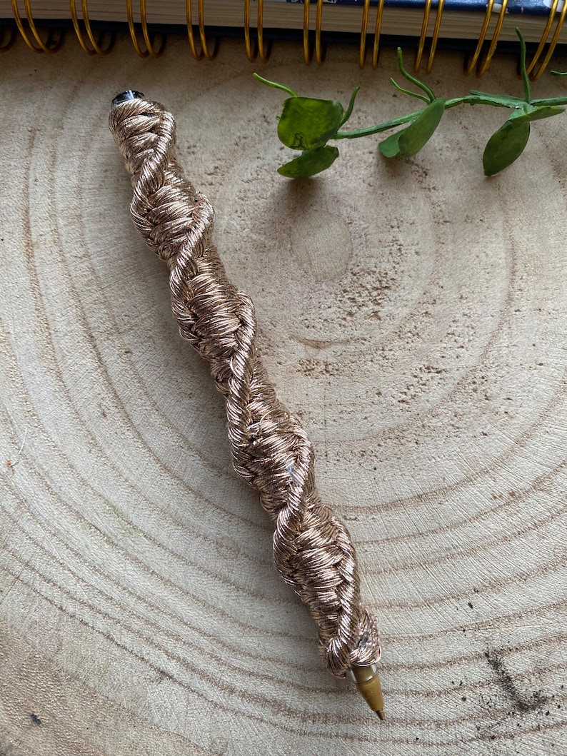 May include: A gold-colored pen with a braided rope design. The pen is lying on a wooden surface.