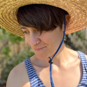 May include: A straw sun hat with a wide brim and a blue chin strap. The hat is made of woven straw and has a round crown. The chin strap is a bright blue cord with a black toggle. The person is wearing a blue and white striped tank top.