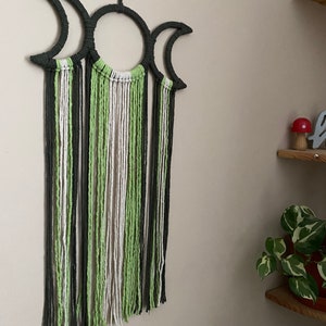 May include: A green, white and black macrame wall hanging with a triple moon design. The hanging has long strands of yarn that create a fringe effect.
