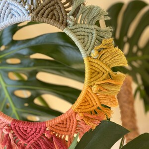 Puede incluir: Un tapiz de macram&eacute; de forma circular con detalles de flecos en colores arco&iacute;ris. El macram&eacute; est&aacute; hecho con diferentes tonos de azul, verde, amarillo y rojo.