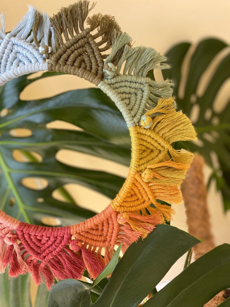 Puede incluir: Un tapiz de macram&eacute; de forma circular con detalles de flecos en colores arco&iacute;ris. El macram&eacute; est&aacute; hecho con diferentes tonos de azul, verde, amarillo y rojo.