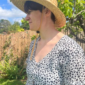 May include: A straw sun hat with a wide brim and a chin strap. The hat is worn with a black and white patterned dress. The background includes a wooden fence, green grass, and a blue sky.