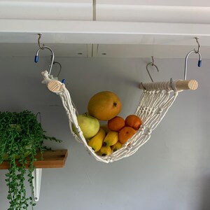 A white macrame hammock hanging from a white cabinet with a wooden dowel rod. The hammock is filled with yellow bananas, oranges and a mango. there is a small houseplant on a shelf nearby.