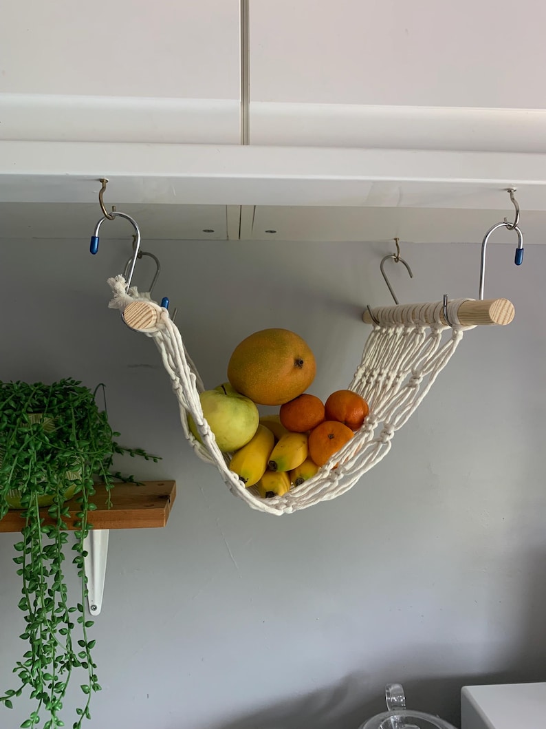 A white macrame hammock hanging from a white cabinet with a wooden dowel rod. The hammock is filled with yellow bananas, oranges and a mango. there is a small houseplant on a shelf nearby.