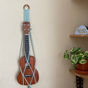 May include: A light blue macrame wall hanging with a wooden ring at the top, holding a brown ukulele. The ukulele is hanging on a white wall.