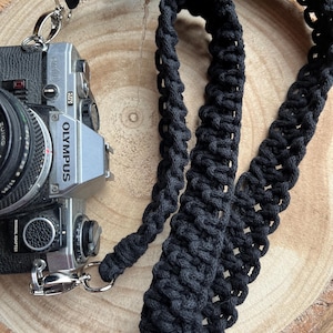 May include: Black camera strap made of braided cotton rope. The strap is attached to a silver camera with the word "OLYMPUS" printed on the side.