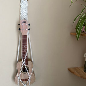 以下が含まれることがあります： 木製リングの上部に白いマクラメの壁掛けがあり、木製のウクレレを保持しています。ウクレレはベージュの壁に掛けられています。