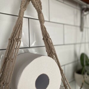Close up detail on handmade macrame toilet paper hammock, made with taupe recycled cotton, hanging under a shelf against a white tiled wall