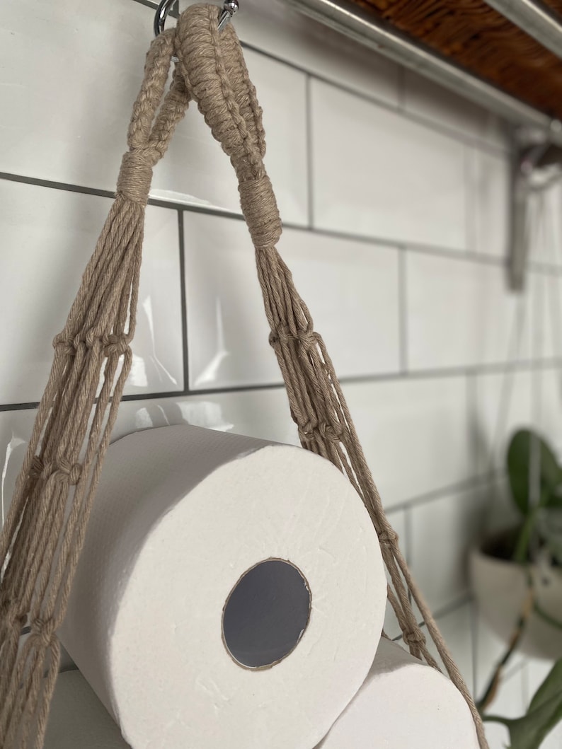 Close up detail on handmade macrame toilet paper hammock, made with taupe recycled cotton, hanging under a shelf against a white tiled wall