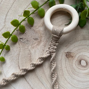 May include: A wooden ring with a natural finish is attached to a beige macrame cord. The cord is intricately knotted in a spiral pattern. Green leafy vines are in the background, creating a natural aesthetic.