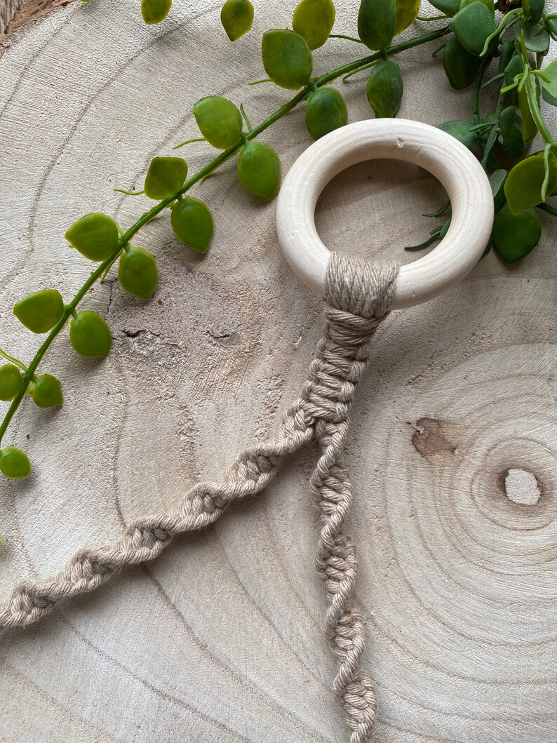 May include: A wooden ring with a natural finish is attached to a beige macrame cord. The cord is intricately knotted in a spiral pattern. Green leafy vines are in the background, creating a natural aesthetic.