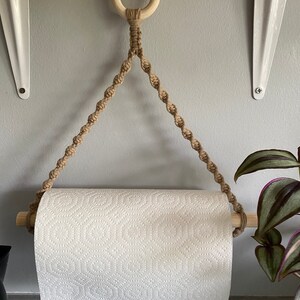 May include: A macrame paper towel holder with a wooden dowel and ring. The holder is made of tan rope and hangs from a wooden shelf. A roll of white paper towels is in the holder.