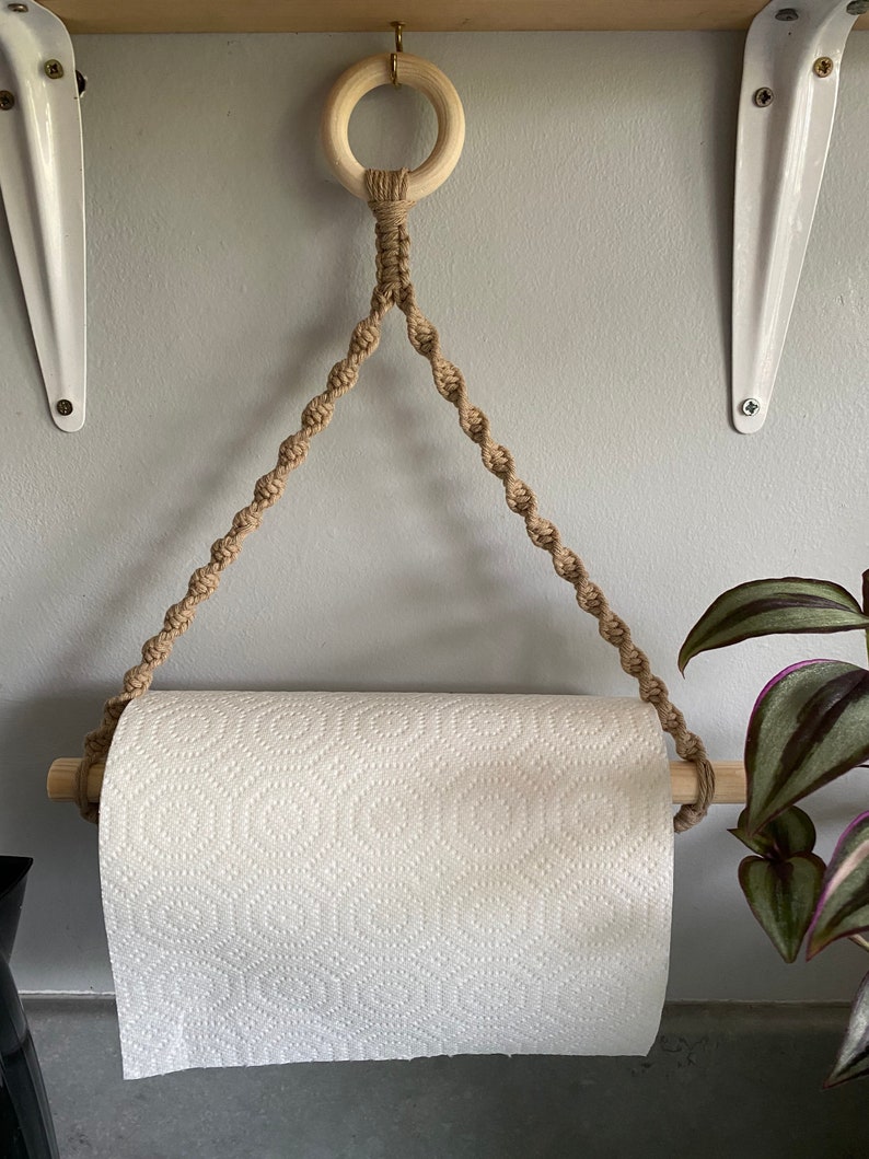 May include: A macrame paper towel holder with a wooden dowel and ring. The holder is made of tan rope and hangs from a wooden shelf. A roll of white paper towels is in the holder.