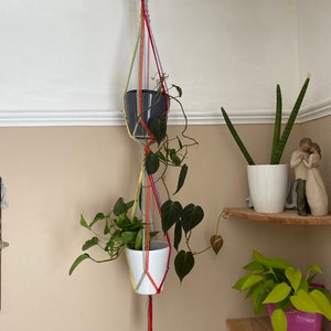 May include: A rainbow macrame plant hanger with two hanging pots. The top pot is grey and the bottom pot is white. The plant hanger is hanging from a wooden ring.