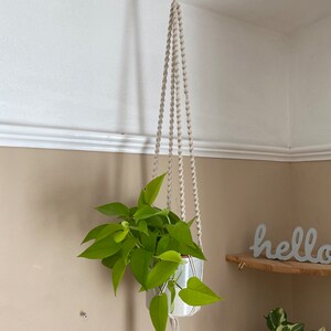 Peut inclure: Un porte-plante en macramé blanc avec une plante verte dans un pot blanc suspendu à un crochet sur un plafond blanc. Le porte-plante a un pompon en bas. Le mot "hello" est écrit sur une étagère en bois à l'arrière-plan.