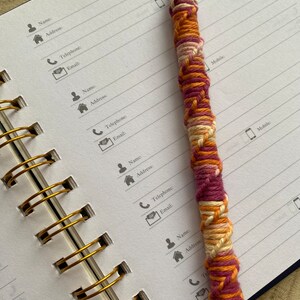 May include: A close-up of a spiral bound notebook with gold rings. The notebook has lined pages and is open to a page with contact information headings. A pen with a pink, orange, and white yarn wrap is lying on the open notebook.