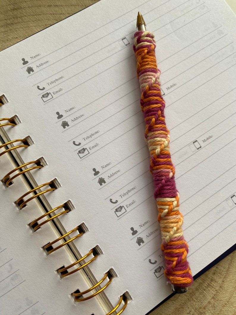 May include: A close-up of a spiral bound notebook with gold rings. The notebook has lined pages and is open to a page with contact information headings. A pen with a pink, orange, and white yarn wrap is lying on the open notebook.