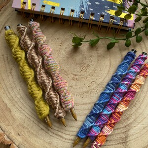 May include: Five colorful yarn-wrapped pens with gold metal tips. The pens are arranged in a fan shape on a wooden surface.