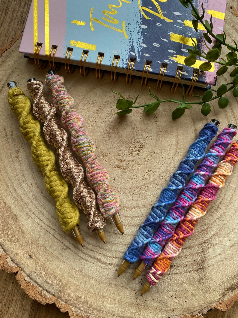 May include: Five colorful yarn-wrapped pens with gold metal tips. The pens are arranged in a fan shape on a wooden surface.