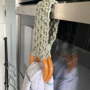Könnte beinhalten: Ein grüner gehäkelter Handtuchring mit zwei Holzringen, die an einem Metall-Ofenhandgriff hängen. Ein weißes Handtuch hängt an den Ringen.
