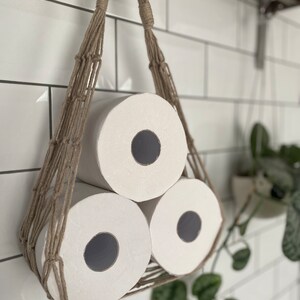 Close up of macrame toilet paper hammock, made with taupe recycled cotton, displayed with 3 large white toilet rolls inside, hanging under a shelf against a white tiled wall