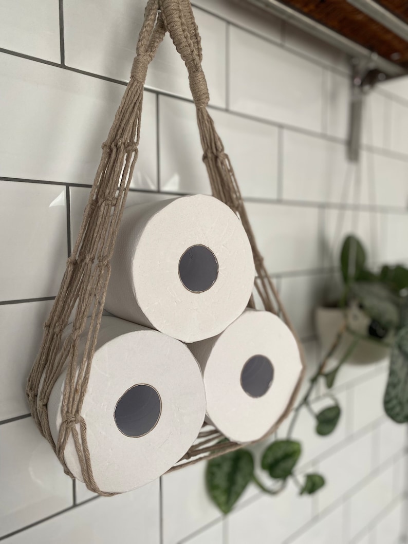 Close up of macrame toilet paper hammock, made with taupe recycled cotton, displayed with 3 large white toilet rolls inside, hanging under a shelf against a white tiled wall