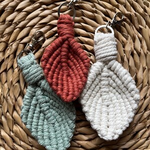 Op de afbeelding: Drie macramé blad sleutelhangers in groen, roest en wit, elk met een zilverkleurige sluiting en sleutelring. De sleutelhangers zijn gerangschikt op een geweven, cirkelvormige placemat, die de handgemaakte textuur en kleuren laat zien.