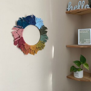 Puede incluir: Un tapiz de macram&eacute; de pared colorido en forma de c&iacute;rculo con un arco&iacute;ris de colores, incluyendo rosa, naranja, amarillo, verde, azul y morado. El tapiz de pared est&aacute; colgado en una pared blanca con un estante de madera encima.