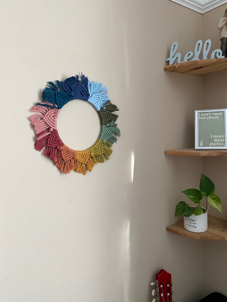 Puede incluir: Un tapiz de macram&eacute; de pared colorido en forma de c&iacute;rculo con un arco&iacute;ris de colores, incluyendo rosa, naranja, amarillo, verde, azul y morado. El tapiz de pared est&aacute; colgado en una pared blanca con un estante de madera encima.