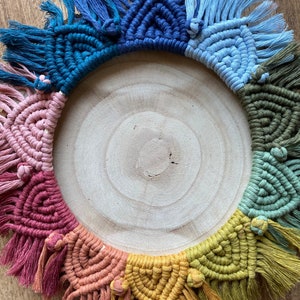 Puede incluir: Un tapiz de macram&eacute; colorido con un arco&iacute;ris de colores, incluyendo rosa, rojo, naranja, amarillo, verde, azul y morado. El macram&eacute; est&aacute; hecho con una variedad de nudos y est&aacute; terminado con borlas. El tapiz se muestra en una rodaja de madera redonda.