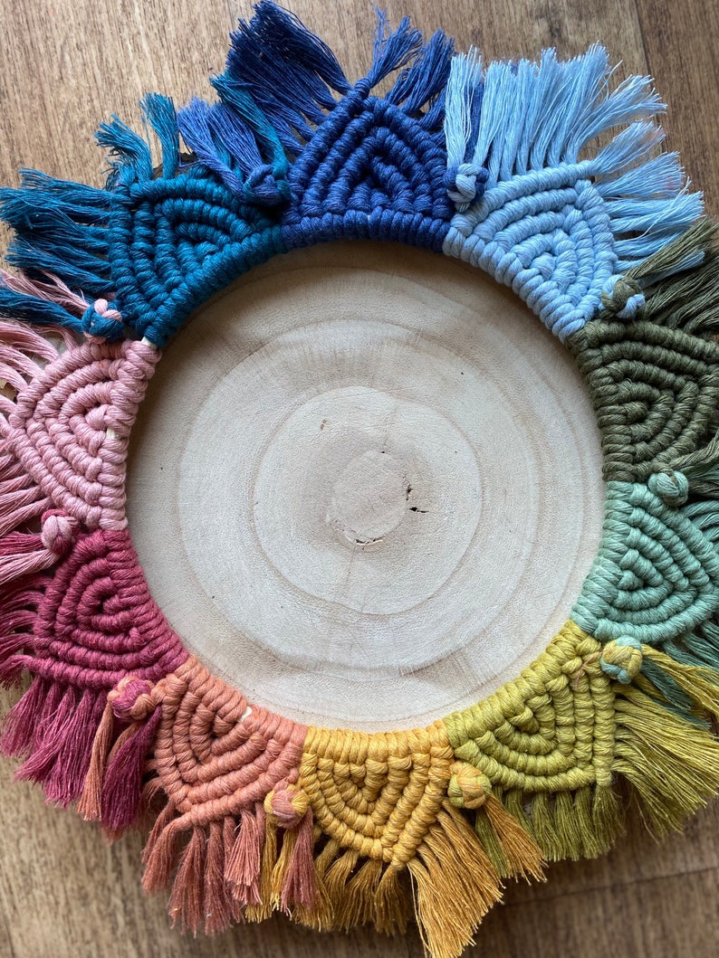 Puede incluir: Un tapiz de macram&eacute; colorido con un arco&iacute;ris de colores, incluyendo rosa, rojo, naranja, amarillo, verde, azul y morado. El macram&eacute; est&aacute; hecho con una variedad de nudos y est&aacute; terminado con borlas. El tapiz se muestra en una rodaja de madera redonda.