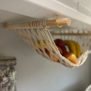 A white string hammock hanging from a wooden dowel rod with a gold hook. The hammock is filled with fruit, including bananas and apples.