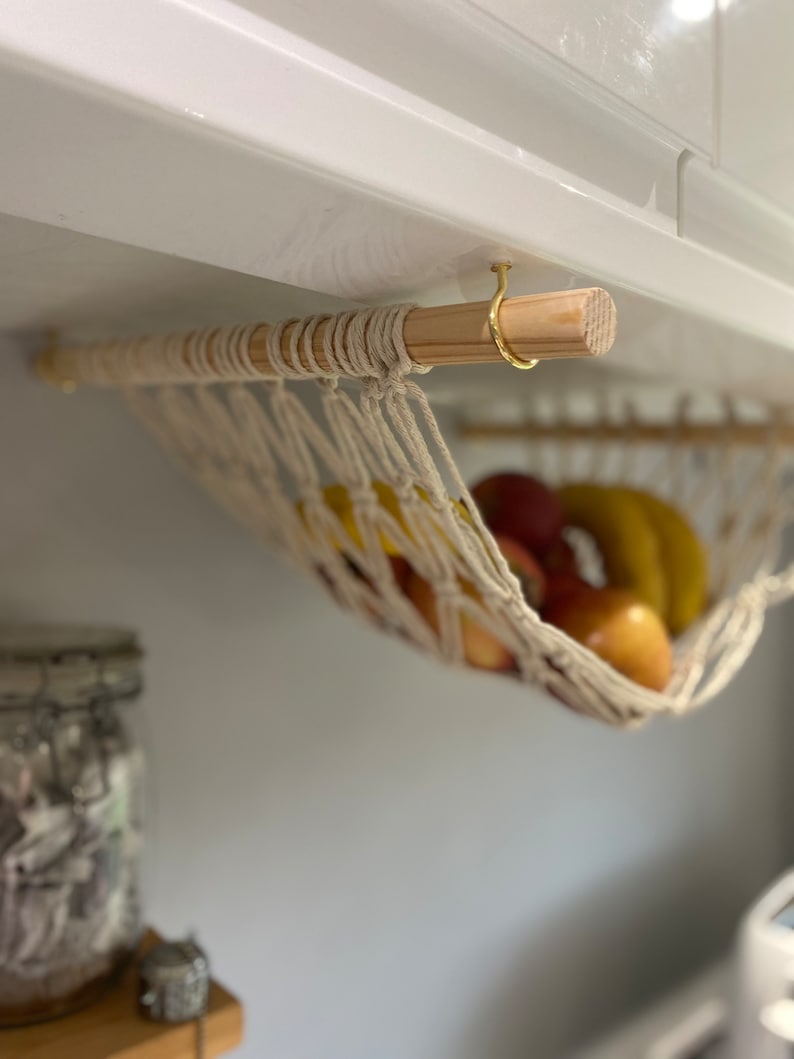 A white string hammock hanging from a wooden dowel rod with a gold hook. The hammock is filled with fruit, including bananas and apples.