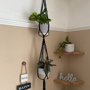 May include: Black macrame plant hanger with two hanging pots. The top pot is gray and the bottom pot is white and beige. The hanger is hanging from a ceiling hook. The pots are filled with green plants. There are wooden signs with the words "Think Happy Be Happy" hanging on the wall.