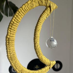 Op de afbeelding: Een gele macramé halve maan wandhanger met een kristallen prisma dat eraan hangt.
