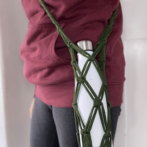 Könnte beinhalten: Ein grüner gehäkelter Wasserflaschenhalter mit einer weißen Wasserflasche im Inneren. Der Halter wird als Umhängetasche getragen.