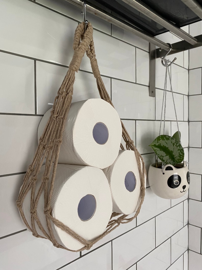 Macrame toilet paper hammock, made with taupe recycled cotton, displayed with 3 large white toilet rolls inside, hanging under a shelf against a white tiled wall
