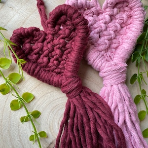 Könnte beinhalten: Zwei handgefertigte Makramee-Herzen mit Quasten als Wandbehänge. Ein Herz ist burgunderrot und das andere rosa.