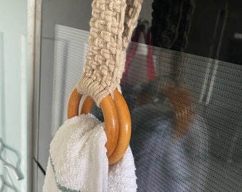 May include: A macrame towel holder with two wooden rings and a white towel with green trim. The macrame is a natural beige color and is looped over a stainless steel appliance handle.