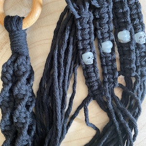 Pode incluir: Um suporte de planta de macramé preto com um anel de madeira no topo e quatro contas de caveira brancas.