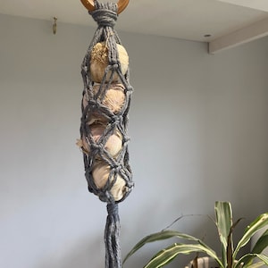 May include: A grey macrame garlic holder with a wooden ring at the top. The holder is filled with garlic bulbs and hangs from a hook.