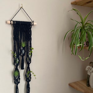 Könnte beinhalten: Schwarze Makramee-Wandbehänge mit drei Glasbehältern für Pflanzen. Die Makramee hängt an einer Holzleiste und einem goldenen Haken. Der Wandbehang befindet sich an einer hellbeigen Wand.