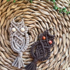 May include: Two macrame owl keychains, one in light beige and one in dark brown, each featuring a metal ring, knotted cord patterns, and two round beads for eyes, with a small tassel at the bottom of each owl figure.
