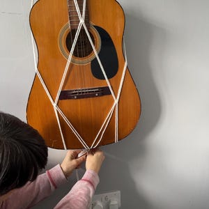 Könnte beinhalten: Eine Person bindet ein weißes Seil an eine braune Akustikgitarre, die an einer Wand hängt. Die Gitarre wird von einer Reihe weißer Seile gehalten, die ein Rautenmuster bilden.