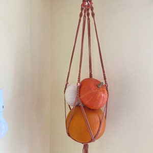 May include: A brown macrame plant hanger with three pumpkins hanging from it. The pumpkins are orange and white.