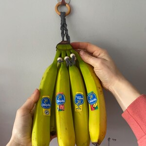 Peut inclure: Un régime de bananes avec des autocollants Chiquita, suspendu à un porte-banane en macramé. Les bananes sont vertes et jaunes, certaines jaunissant davantage. Le support a un anneau en bois et une corde grise.