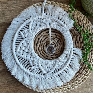 May include: A white macrame wall hanging with a crystal hanging in the center. The macrame is in the shape of a crescent moon with fringe around the edges.