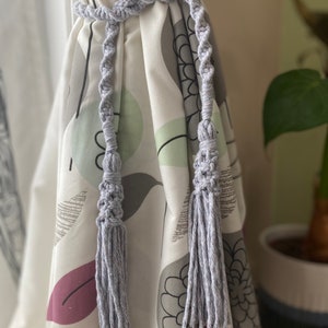 Può includere: Un fermatenda in macramè grigio chiaro con un design intrecciato e estremità a nappa. Il fermatenda è avvolto attorno a una tenda fantasia con disegni floreali bianchi, grigi e viola. L'estetica generale è bohémien e decorativa.
