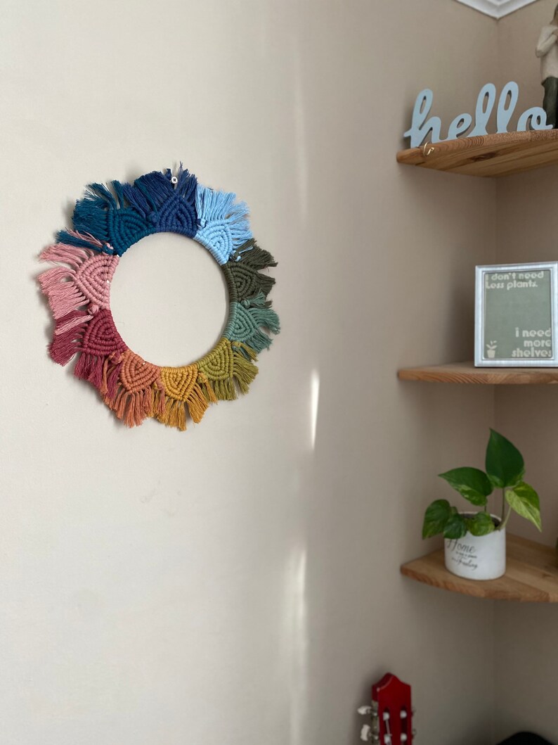 Puede incluir: Un tapiz de macram&eacute; de pared colorido con forma circular y flecos. El tapiz est&aacute; hecho con diferentes tonos de rosa, rojo, naranja, amarillo, verde, azul y azul marino.