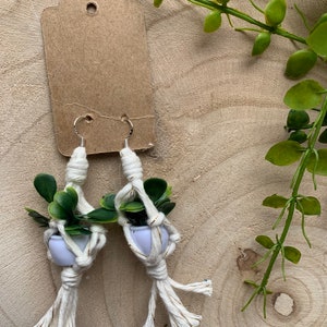 May include: A pair of white macrame earrings with small green plants in white pots. The earrings are hanging from silver hooks.